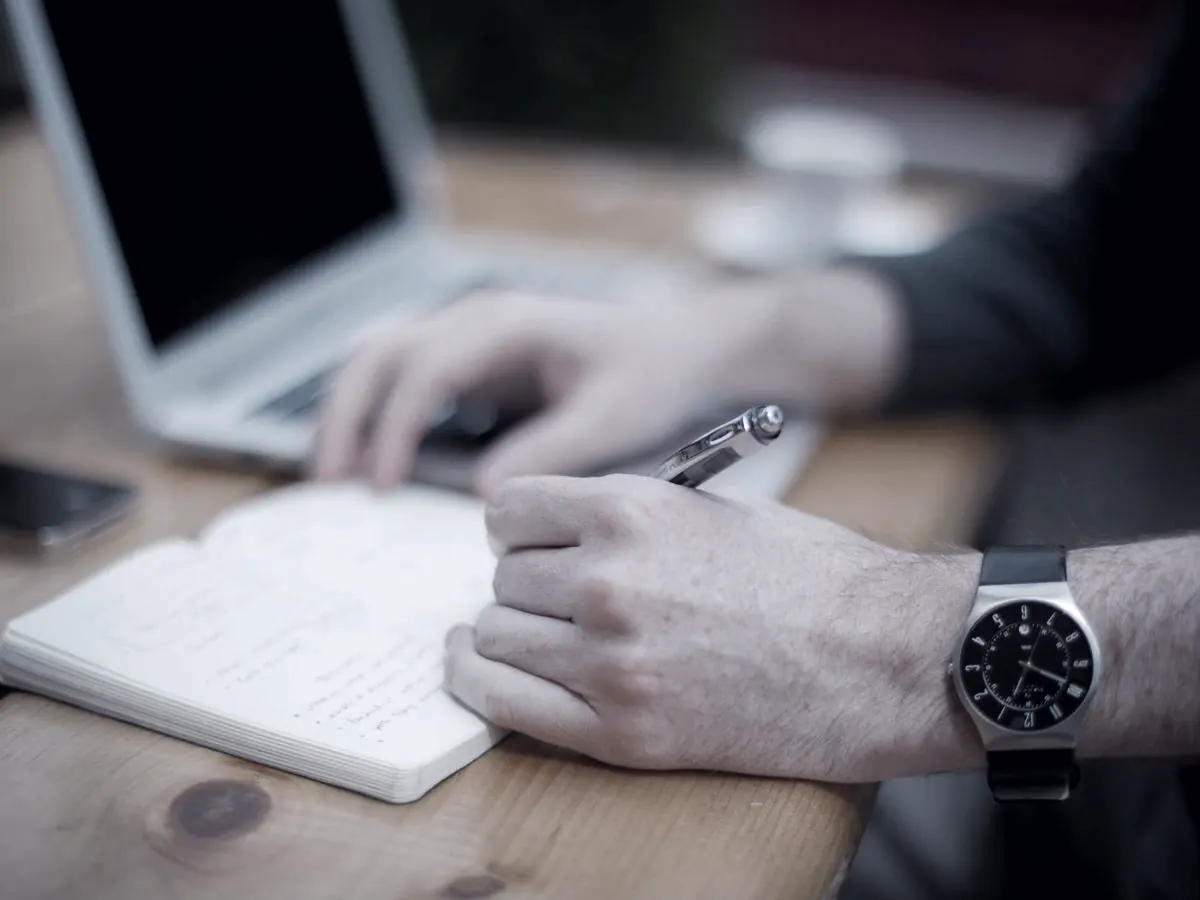 Hands writing in a notebook beside a laptop
